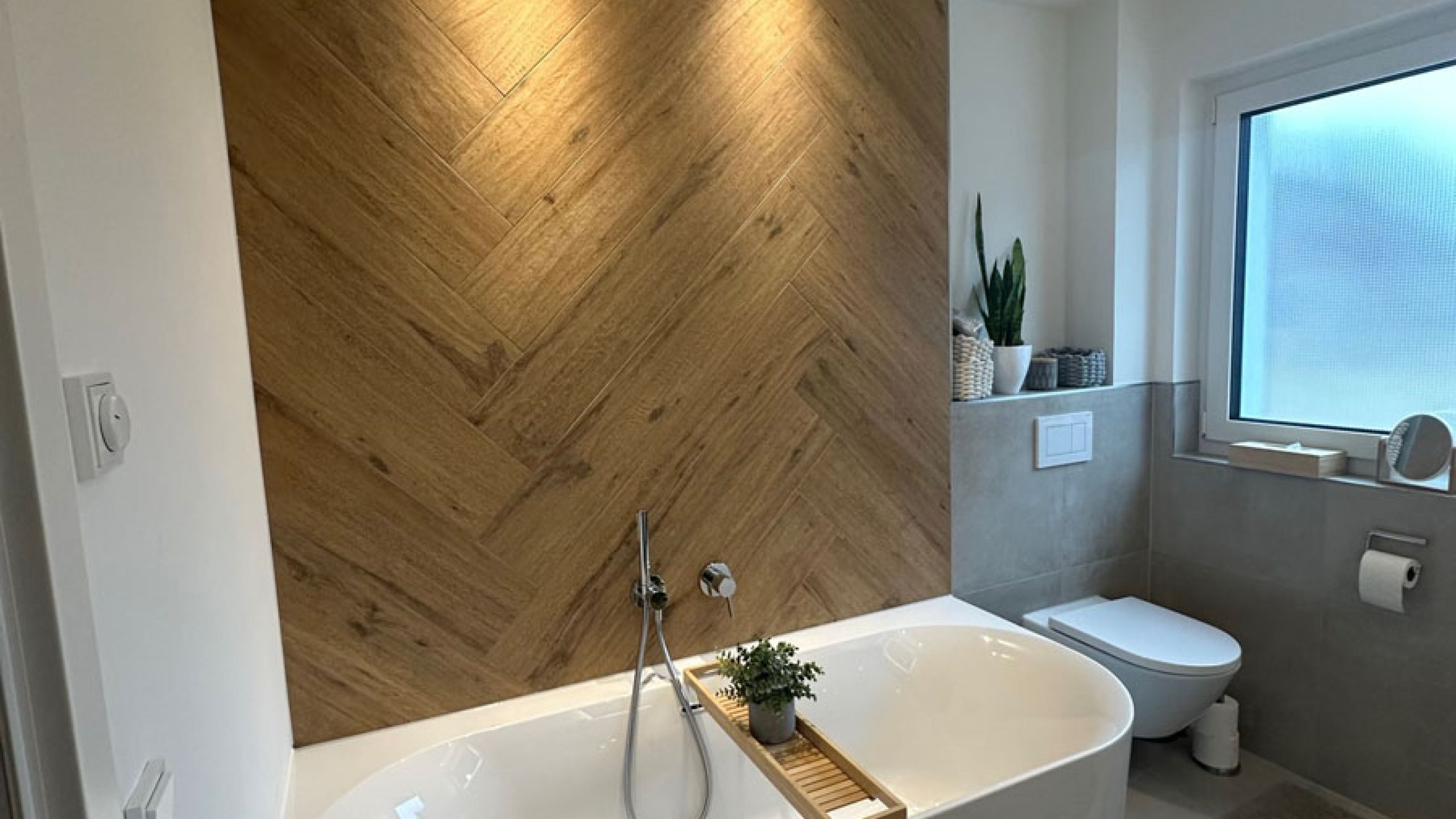 Badezimmer mit Badewanne vor Fischgrät-Wand in Holzoptik, grauen Wandfliesen und Wand-WC