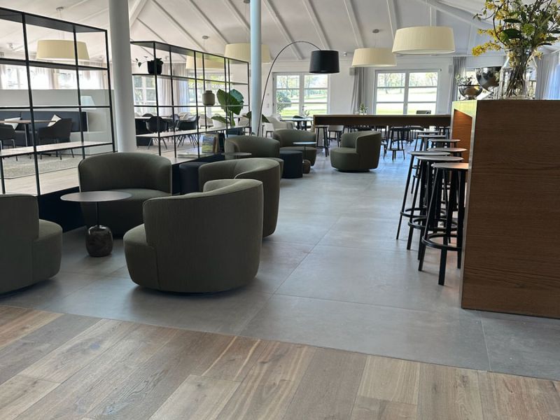 Lounge mit hellgrauen Großformatfliesen in Betonoptik, Holzboden im Vordergrund, Sesseln und Bartheke