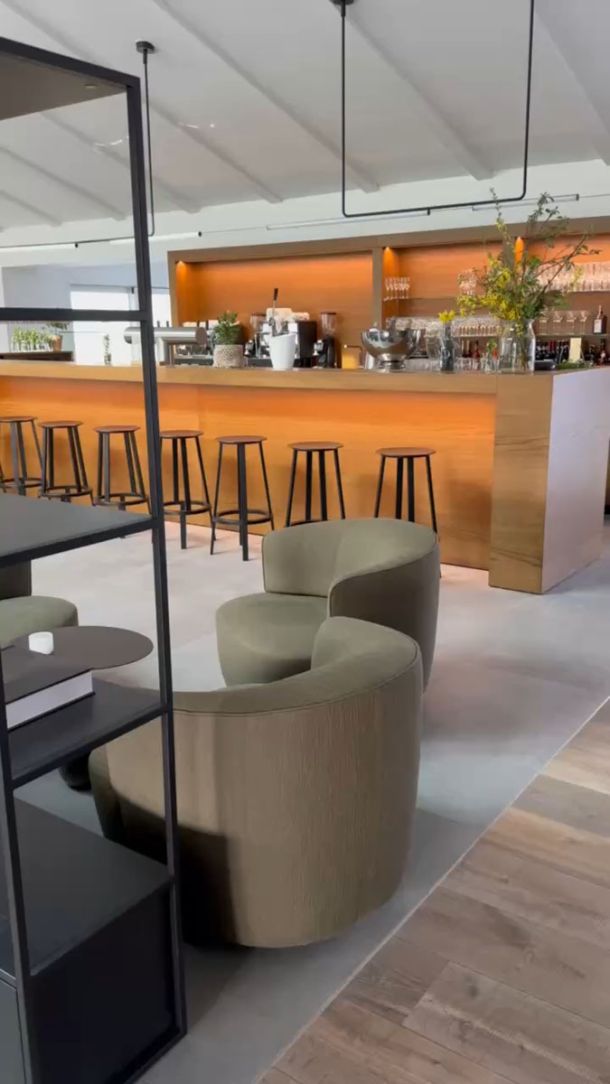 Bartheke mit Holzfront und Barhockern, davor Sessel auf hellgrauen Großformatfliesen in Betonoptik
