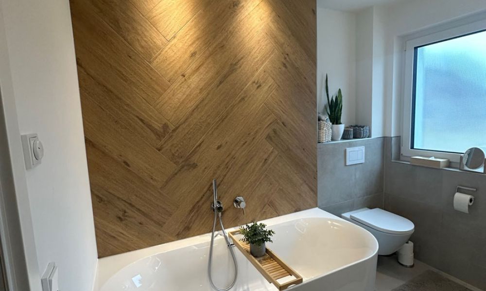 Badezimmer mit Badewanne vor Fischgrät-Wand in Holzoptik, grauen Wandfliesen und Wand-WC