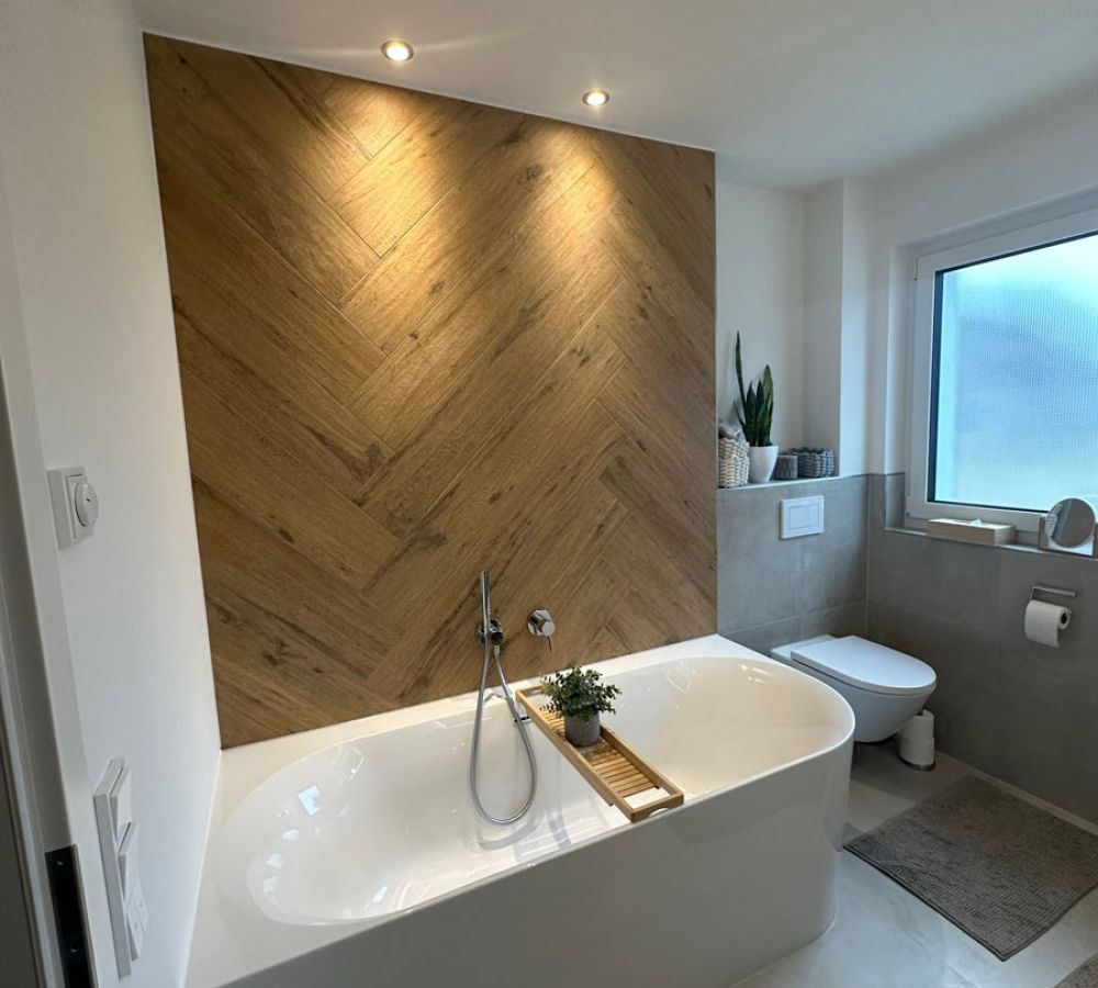Badezimmer mit Badewanne vor Fischgrät-Wand in Holzoptik, grauen Wandfliesen und Wand-WC