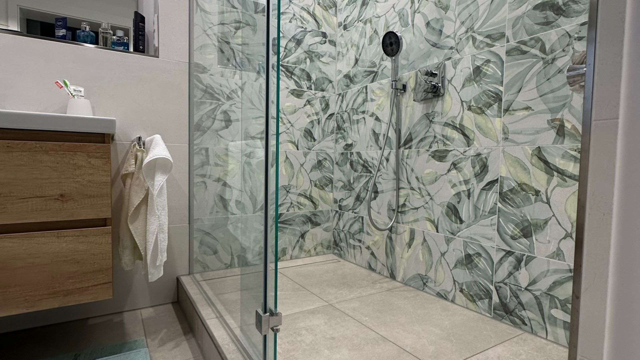 Dusche mit Glastür, Wandfliesen mit grünem Blätterdekor und Bodenfliesen in heller Natursteinoptik